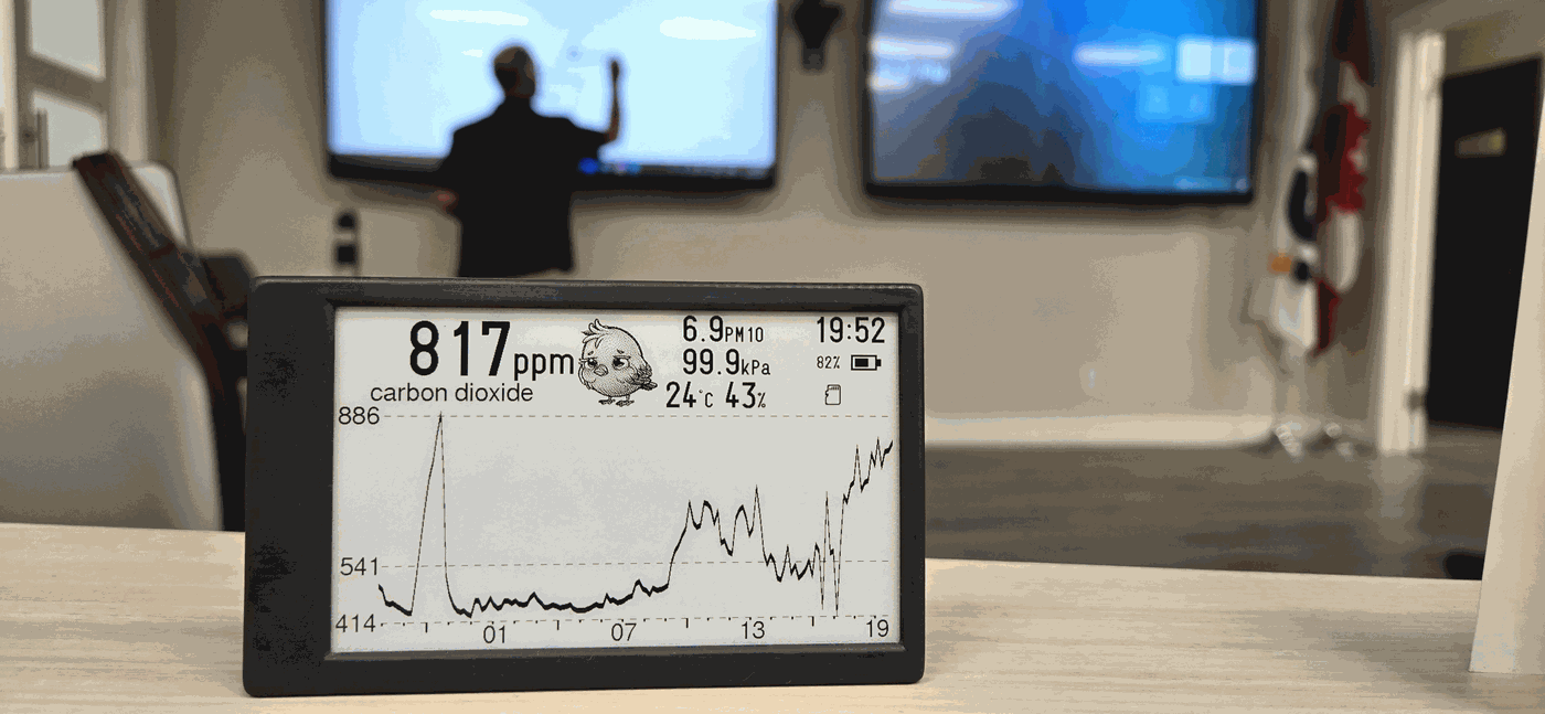 A Canary on a boardroom table reading 817 ppm carbon dioxide, its 24-hour curve flat overnight and climbing steadily through the afternoon, with a presentation running on screens behind it.