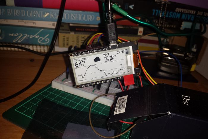 A bare e-paper module and CO2 sensor wired to a breadboard on a desk in front of a stack of books, showing 647 ppm.