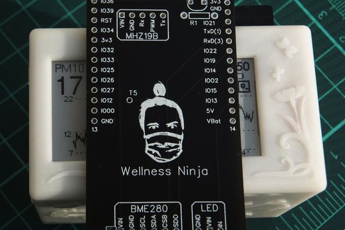 A custom circuit board silkscreened with a line portrait of its maker, Constantin Bucataru, wearing a face mask, above the words Wellness Ninja. It rests against two white resin enclosures.