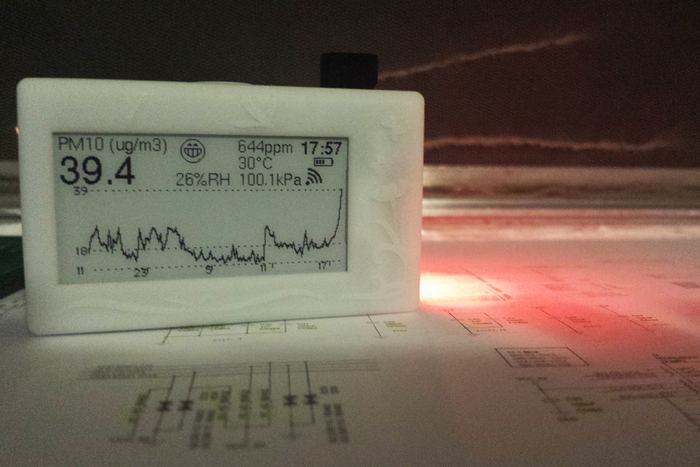 A white resin unit on a desk of schematics at night, its particulate trace climbing to 39.4 PM10 under an orange smoke-lit sky.