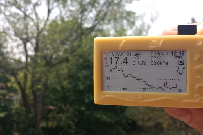 A yellow-cased unit held up against summer trees, showing 117.4 PM10 after a steep climb.