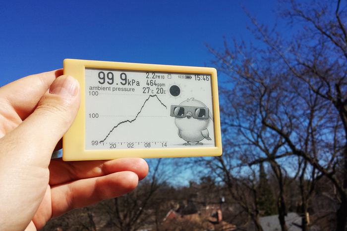 A yellow-cased unit held against a clear blue sky, showing ambient pressure peaking, with the Canary bird wearing eclipse glasses on screen.